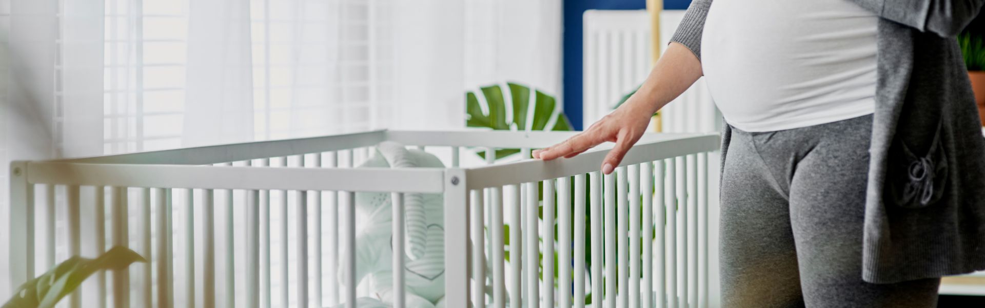 A pregnant woman rests her hand on the rail of a white wooden baby crib in a sunlit nursery.