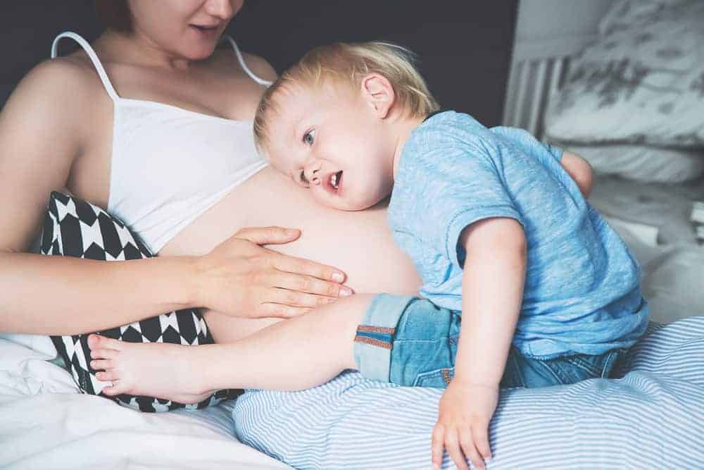 A toddler rests his head on his pregnant mother's bare belly while sitting on a bed.