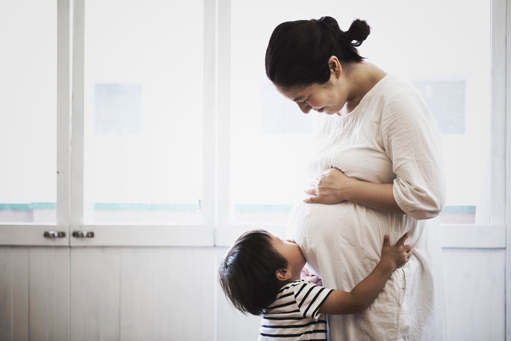 A young boy in a striped shirt hugs and kisses his pregnant mother's belly in front of a bright window.