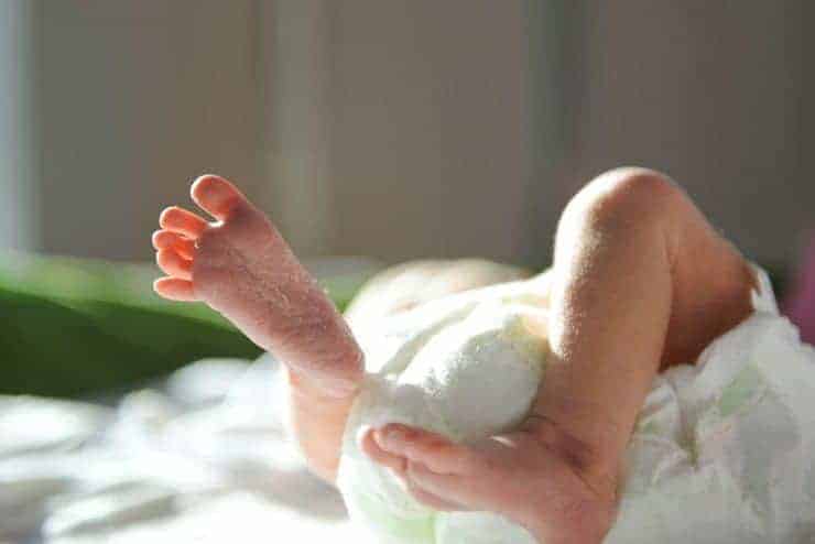 A preterm baby lies on a softly lit bed with tiny feet raised and curled legs, reflecting the early days of neonatal care and recovery under warm light.