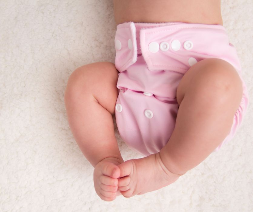 A baby wearing a pink reusable cloth diaper with white snaps lying on a white textured blanket.