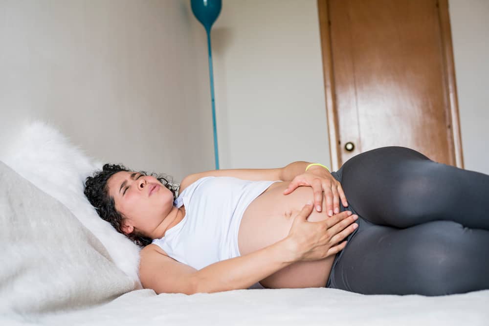Pregnant person lying on a bed with an expression of pain while holding their lower abdomen and pubic area.
