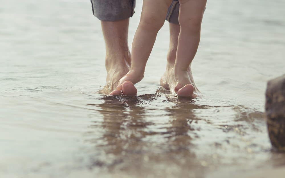 A toddler’s bare feet stepping on an adult’s feet while standing in shallow water.
