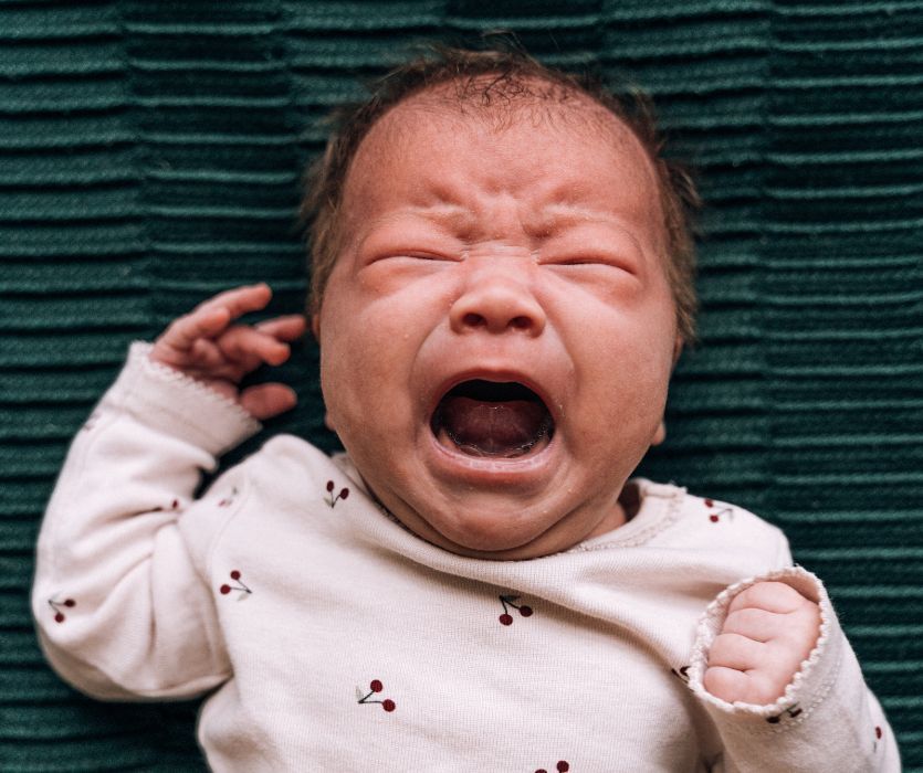 A newborn baby with eyes tightly shut and mouth wide open, crying while lying on a dark green textured surface.
