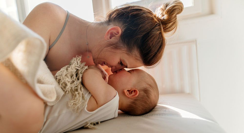 A smiling woman leans over a baby lying on a bed, touching her nose to the baby's nose.