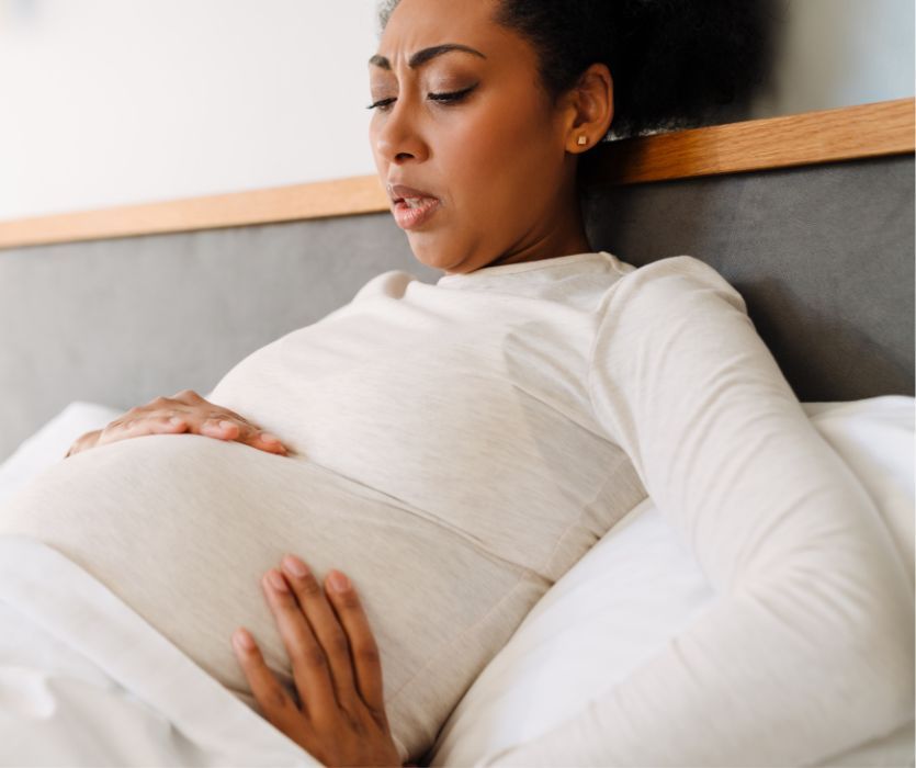 Pregnant Black woman in a bed winces in pain with her hands on her stomach.