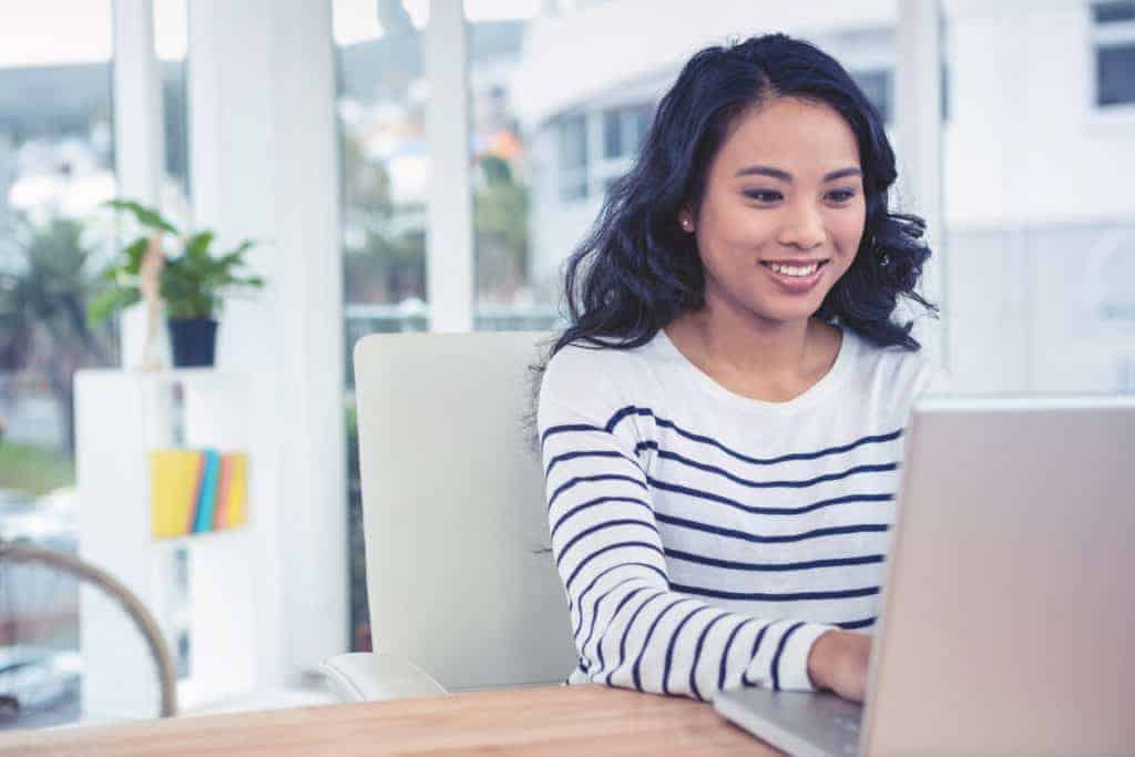Smiling woman in a striped shirt sitting at a desk and using a laptop in a bright office.