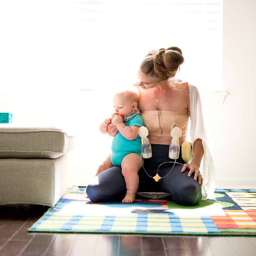 Woman wearing a hands-free pumping bra while holding her baby on a colorful play mat.