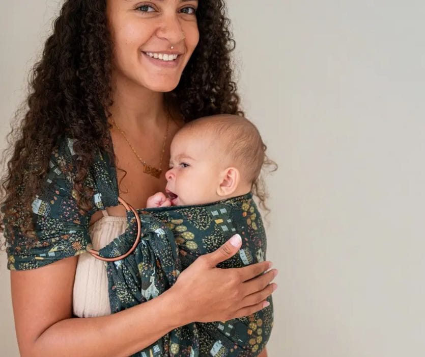 A smiling woman carries an infant in a dark green patterned ring sling.