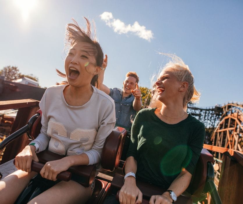 Three people smiling and screaming while riding a wooden roller coaster on a sunny day.