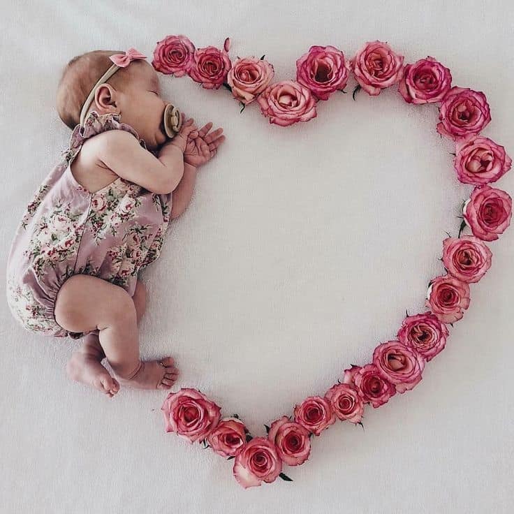 Sleeping newborn in a floral romper lying next to a heart shape made of pink roses on a white background.