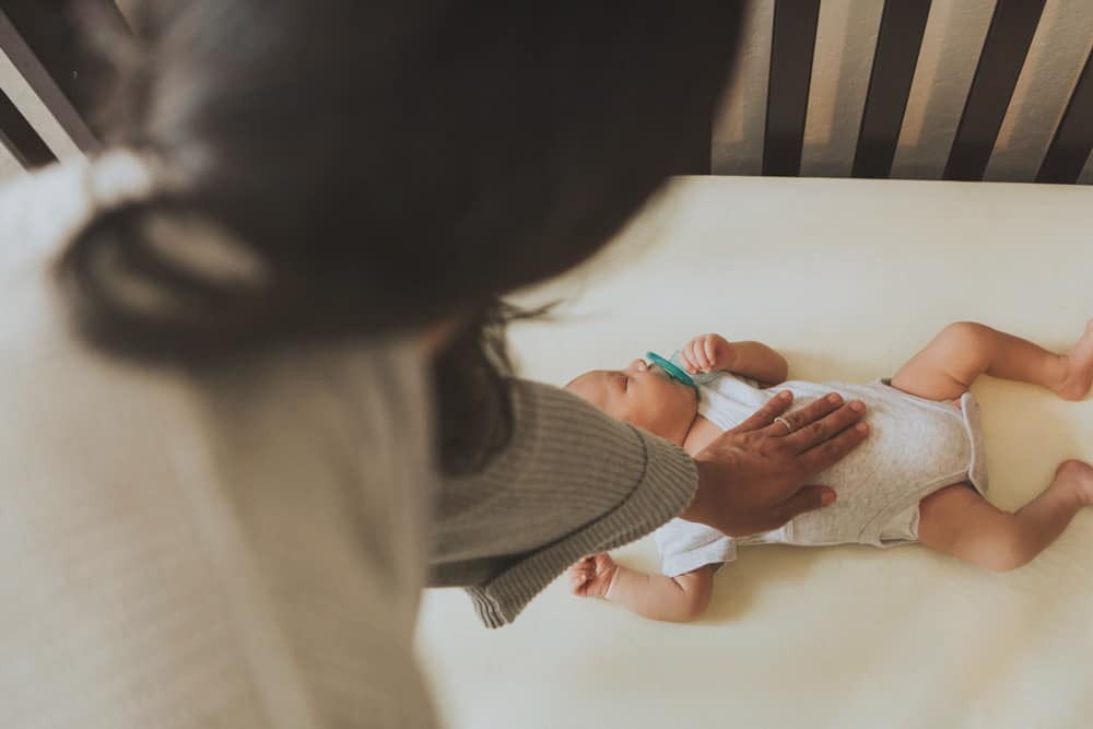 A person rests a hand on a sleeping baby lying on their back in a crib.