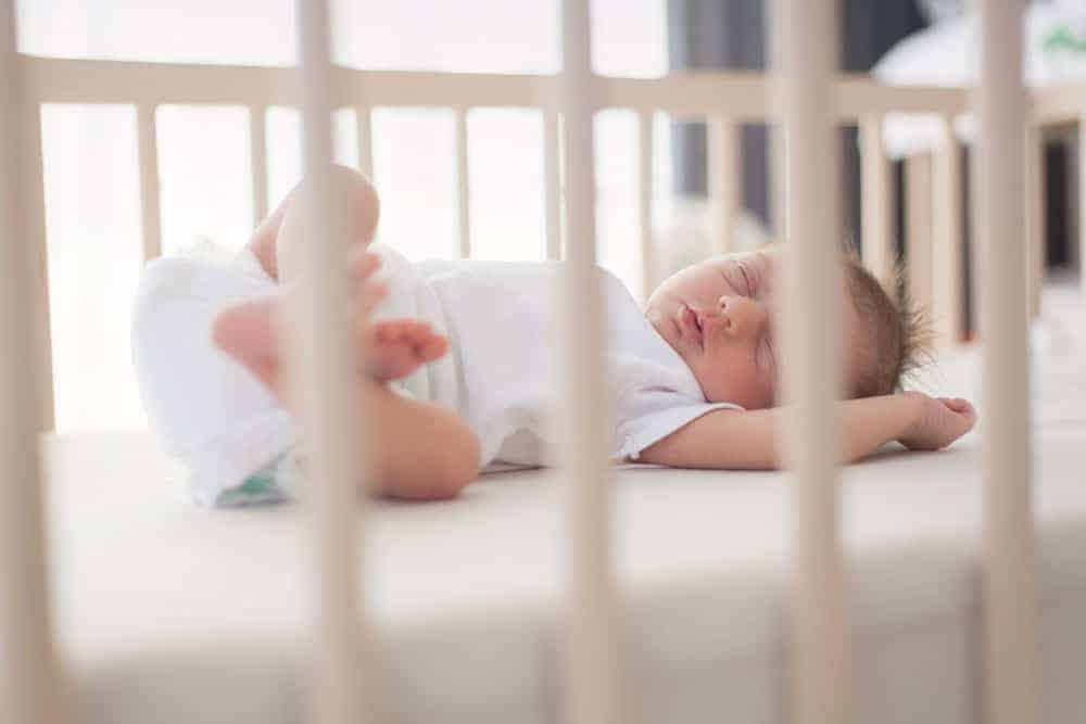 A baby sleeping on their back in a wooden crib with a firm, flat mattress and no loose bedding.