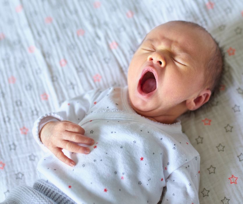 Newborn baby in a star-patterned onesie yawning while lying on a flat, star-printed sheet.