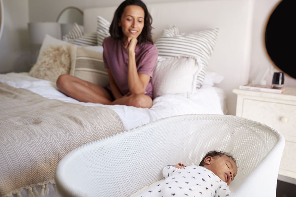 A woman sits on a bed watching a baby sleep on their back in a mesh bassinet next to the bed.