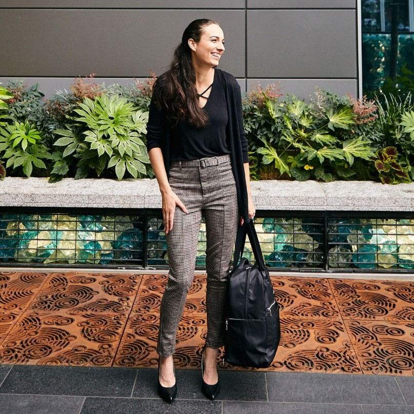 Smiling woman in plaid pants and a black top standing outdoors, holding a large black Sarah Wells breast pump bag.
