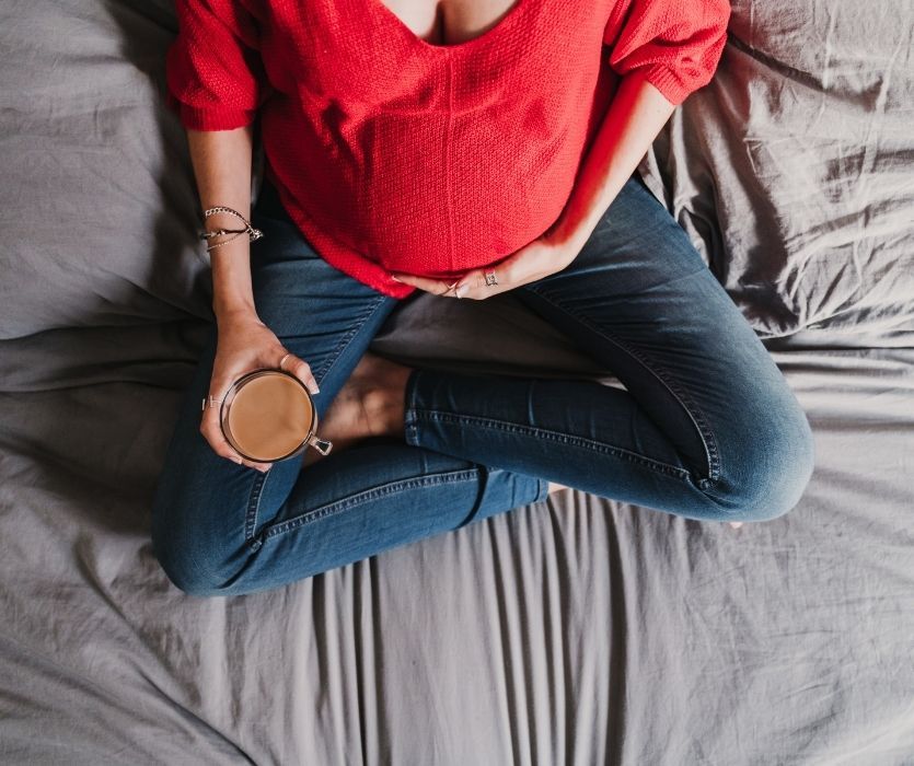 A pregnant woman in a red sweater and jeans sits cross-legged on a bed, holding a glass mug of coffee.
