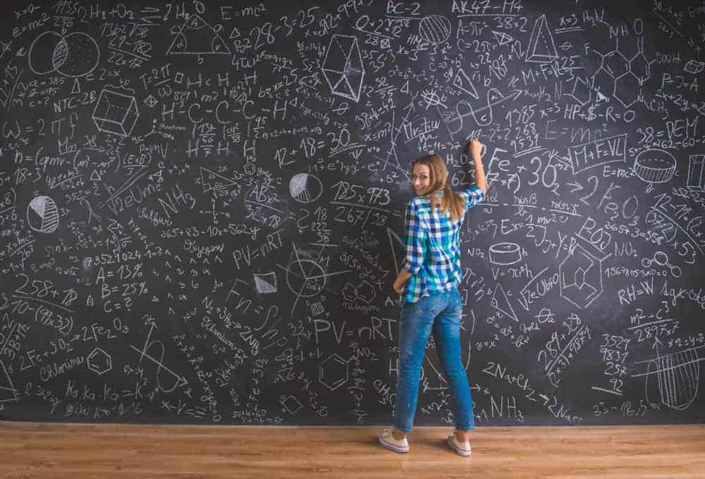 Woman in a plaid shirt writing on a large blackboard covered in complex mathematical and scientific equations.