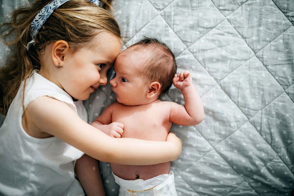 A young girl lies on a quilted blanket and smiles while hugging her newborn sibling.