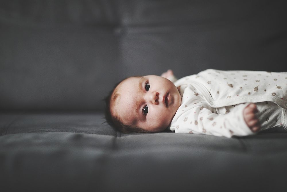 A newborn baby wearing a patterned onesie lies on a dark gray tufted surface.
