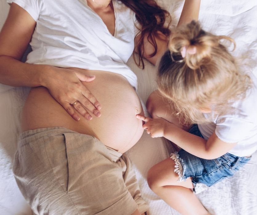 Young girl pointing at her mother's pregnant belly while they lie on a white bed.