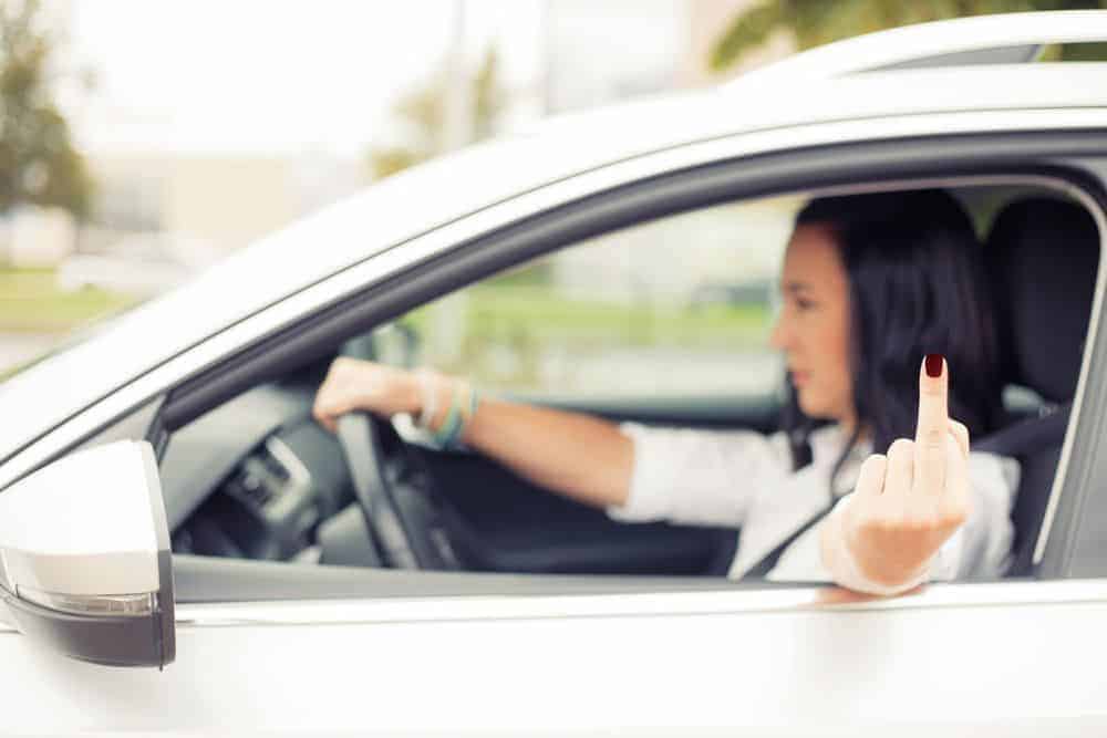A woman driving a white car extends her middle finger out of the window in an aggressive gesture.
