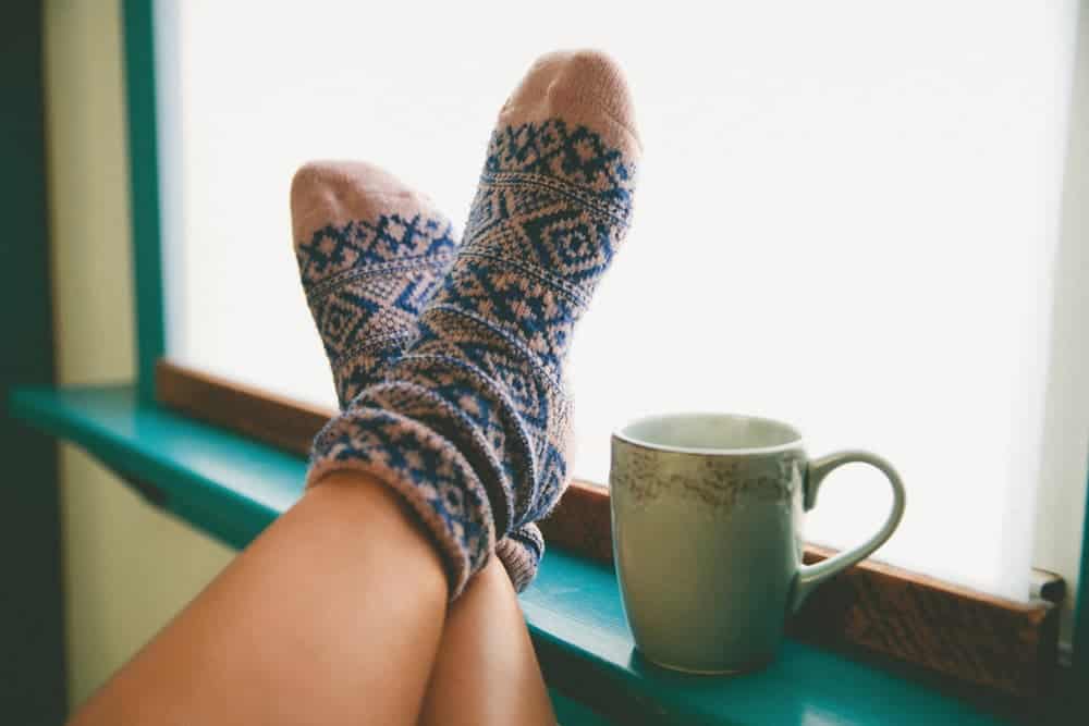 Feet in patterned wool socks resting on a window sill next to a ceramic mug.