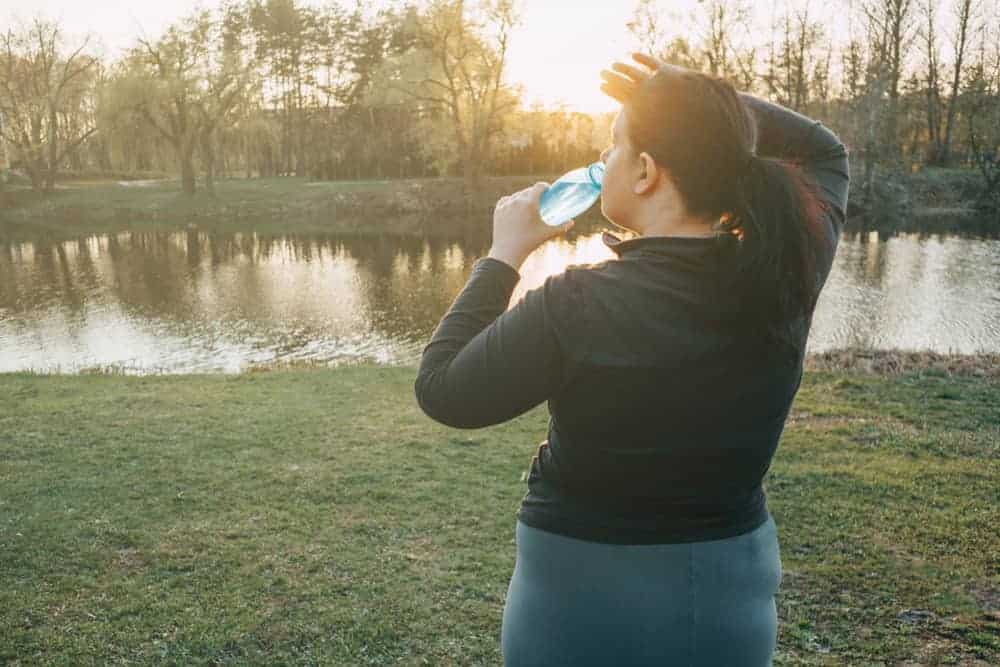 A woman in athletic wear drinks from a water bottle while resting by a lake at sunset.