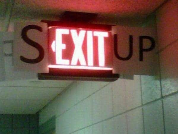 Illuminated red exit sign with paper letters S and UP taped to the sides, making it spell SEX IT UP.