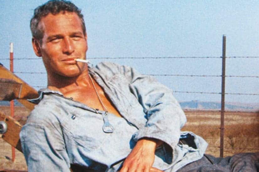 Paul Newman as Cool Hand Luke leans back with a cigarette in his mouth, wearing a blue shirt and bottle opener necklace.