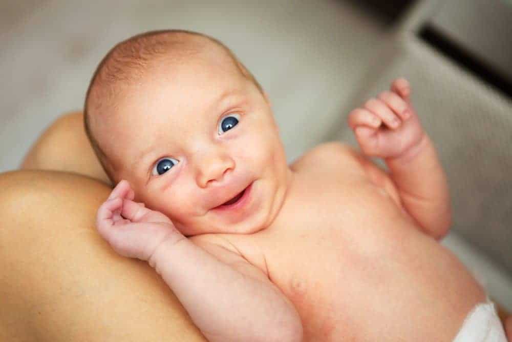 A smiling newborn baby with blue eyes lies on a person’s lap and looks toward the camera.