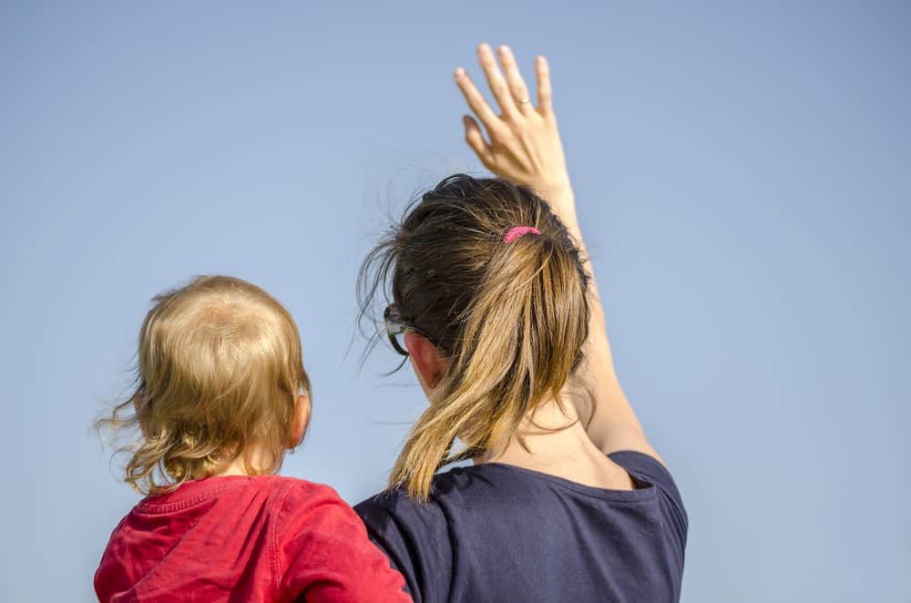 Back view of a woman holding a young child and waving her hand toward a clear blue sky.