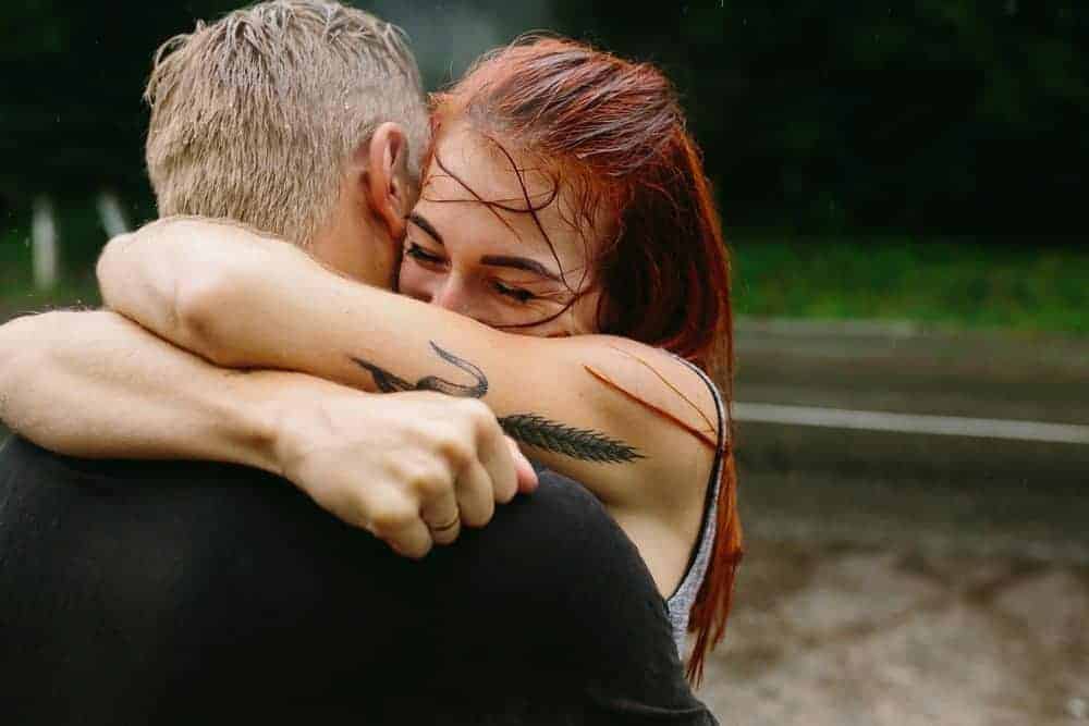 A woman with wet red hair and an arm tattoo smiles while hugging a man in the rain.