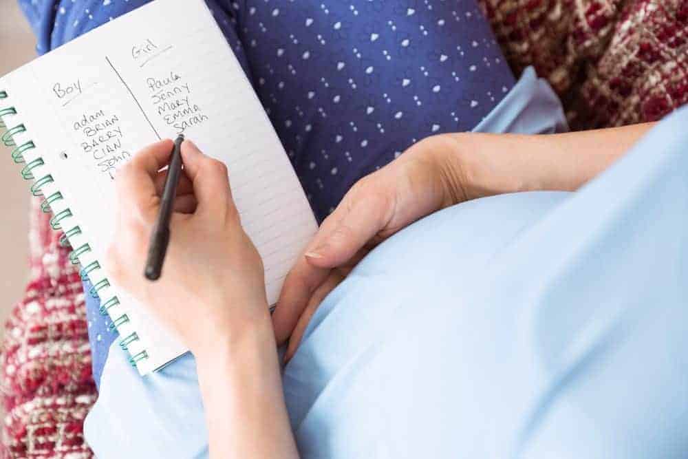 A pregnant woman holds a pen over a notebook listing potential baby names for boys and girls.