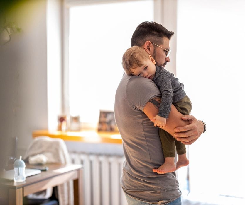 A man holding a tired toddler on his shoulder while standing in a bright room.
