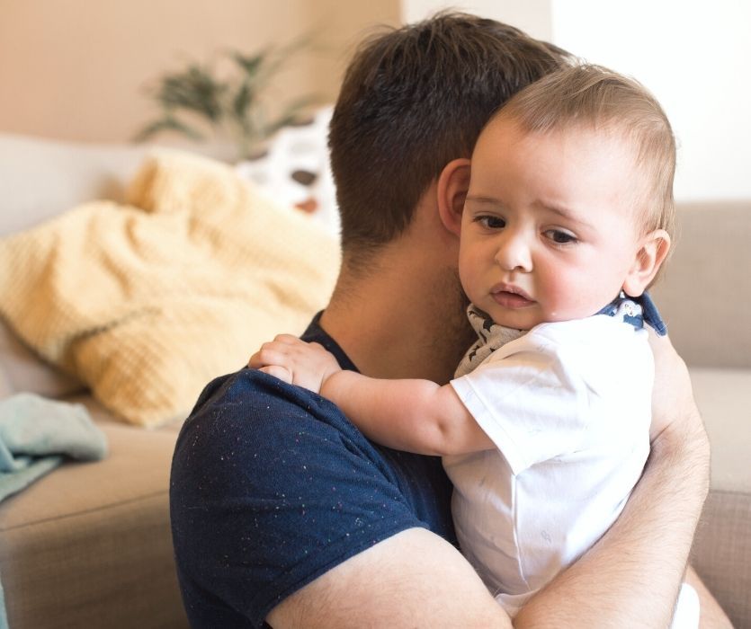 A man holding a sad-looking baby with a white shirt and bib over his shoulder.