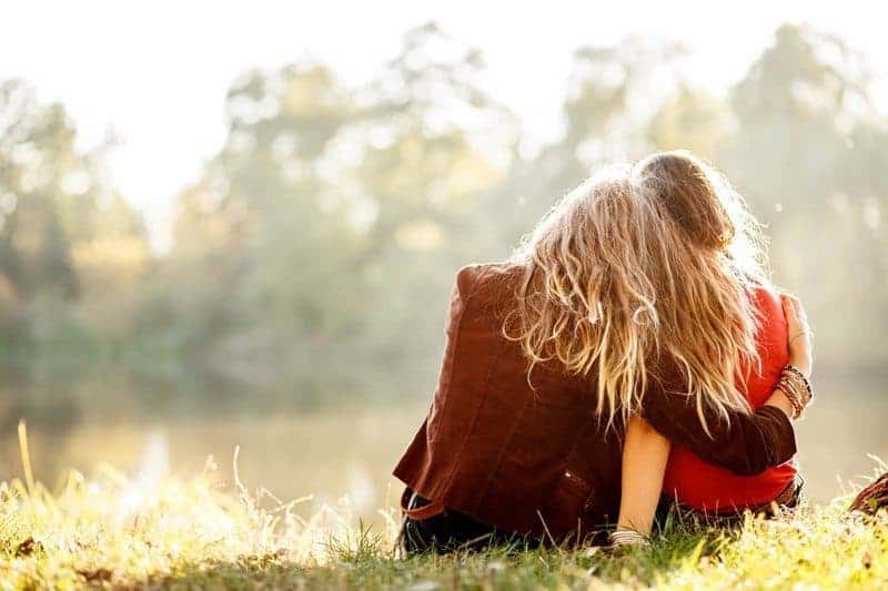Two sisters sitting by a lake, seen from behind, as one rests her head on the other's shoulder in the sunlight.