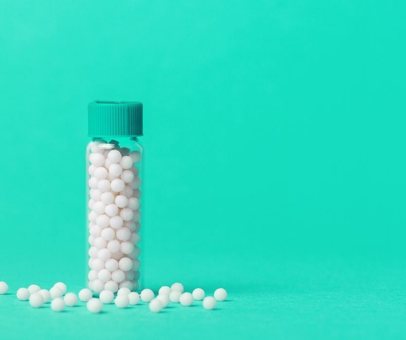 A clear glass vial with a teal cap filled with small white homeopathic globules against a solid teal background.