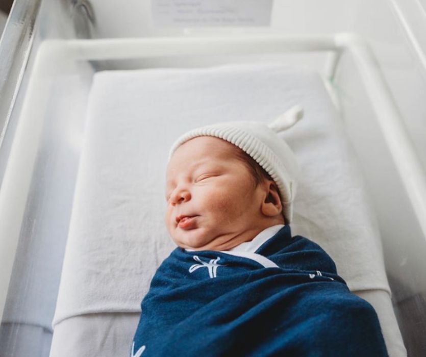 A sleeping newborn baby wearing a white knit hat and wrapped in a blue swaddle in a hospital bassinet.