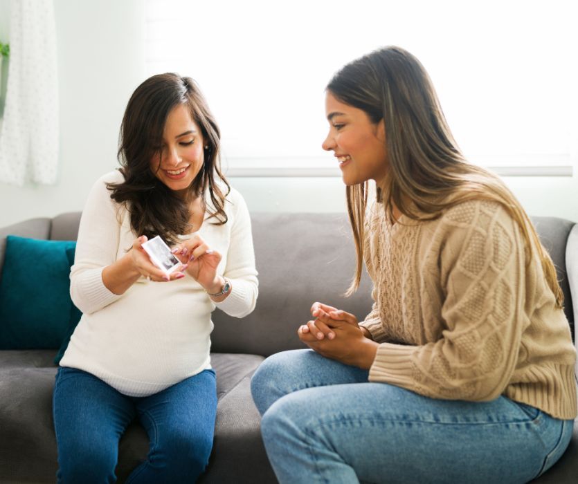 Two smiling women sit on a sofa as one shows the other a sonogram photo.