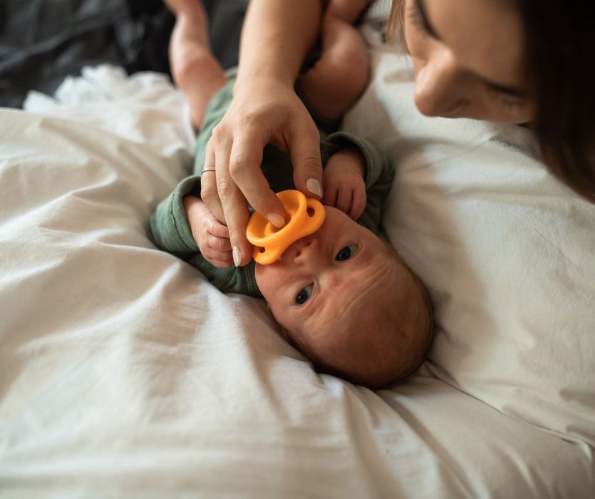 An adult places an orange pacifier into the mouth of a newborn baby lying on a white bed.