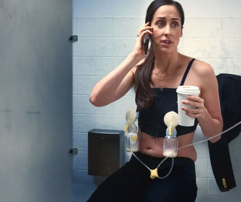 A woman in a bathroom stall using a hands-free breast pump while talking on a cell phone and holding a coffee cup.