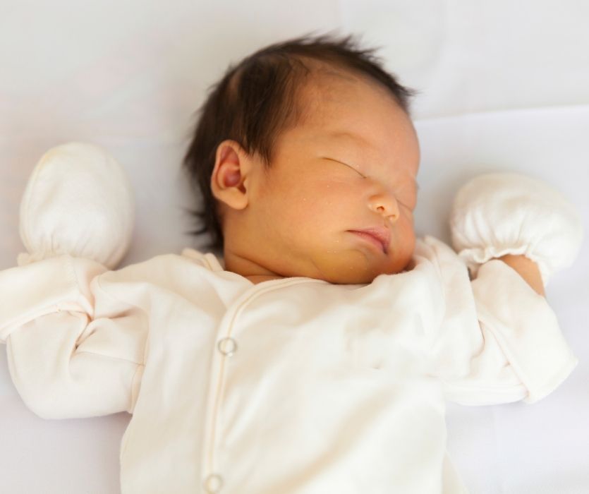 Newborn baby with dark hair sleeping on a white surface, wearing white mittens and a white onesie.