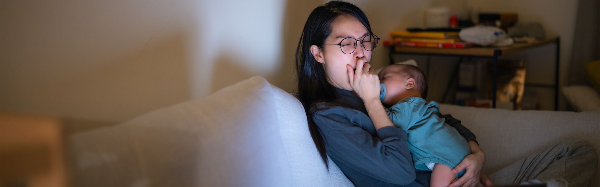 A tired mother with glasses cradles a sleeping baby on a sofa, covering her mouth as she struggles to stay awake.