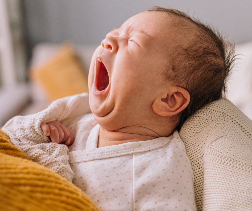 Close-up of a yawning newborn infant resting on a white cable knit blanket.