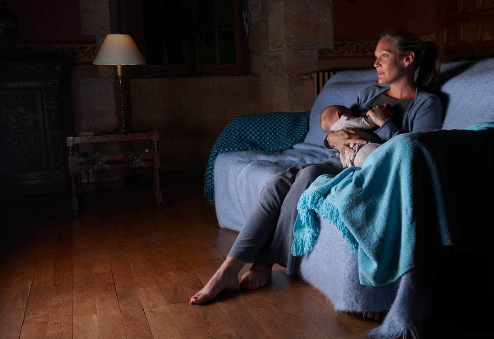 A mother sits on a sofa in a dimly lit room, holding and nursing her sleeping baby at night.