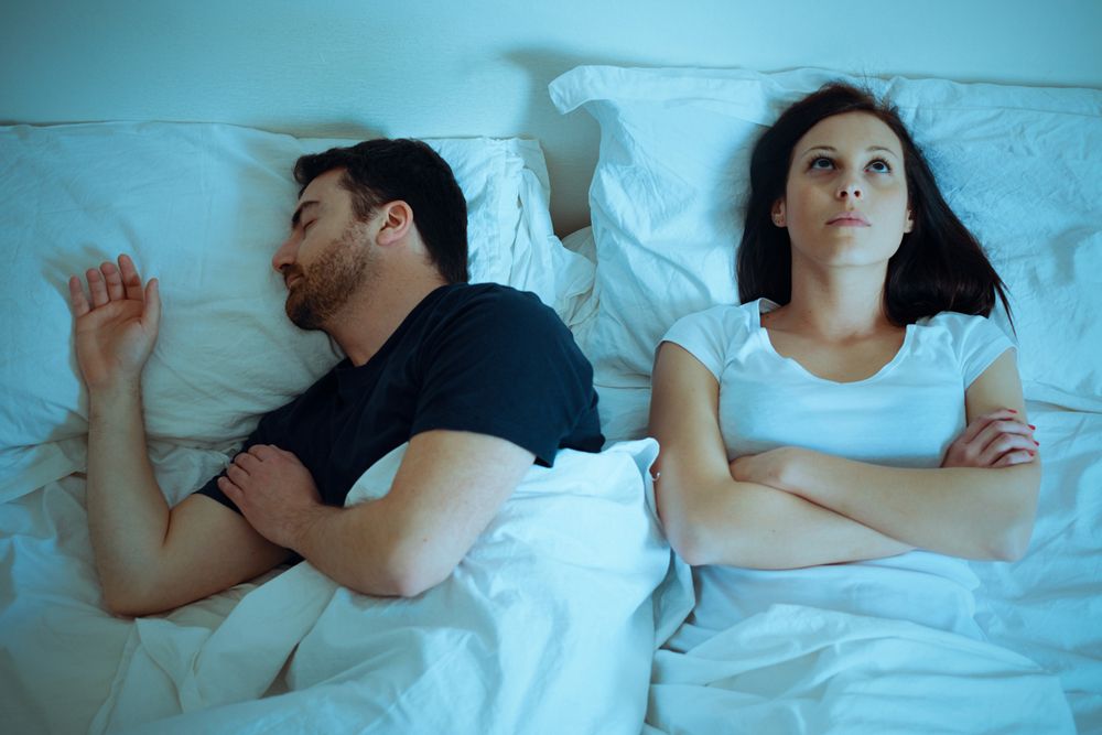 A man sleeps soundly in bed while a woman lies awake next to him with her arms crossed, looking frustrated.