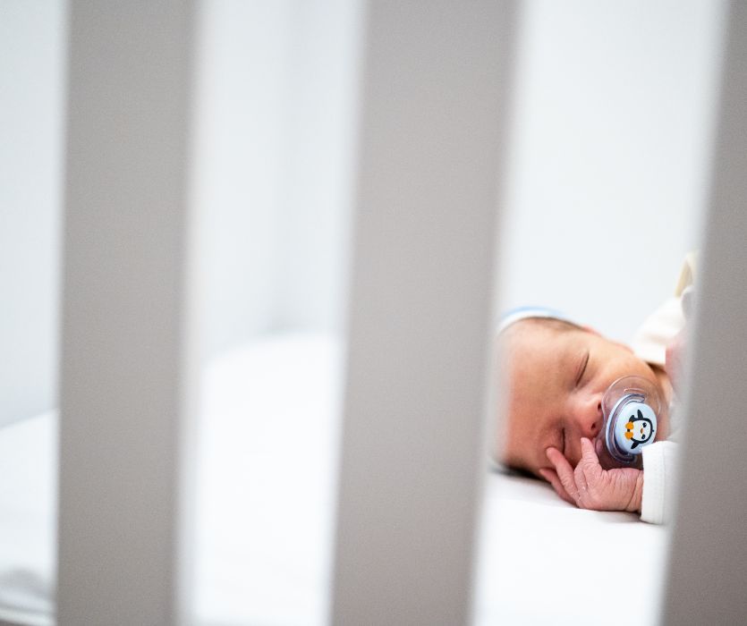 Close-up of a sleeping newborn baby in a white crib with a penguin-patterned pacifier, viewed through crib slats.