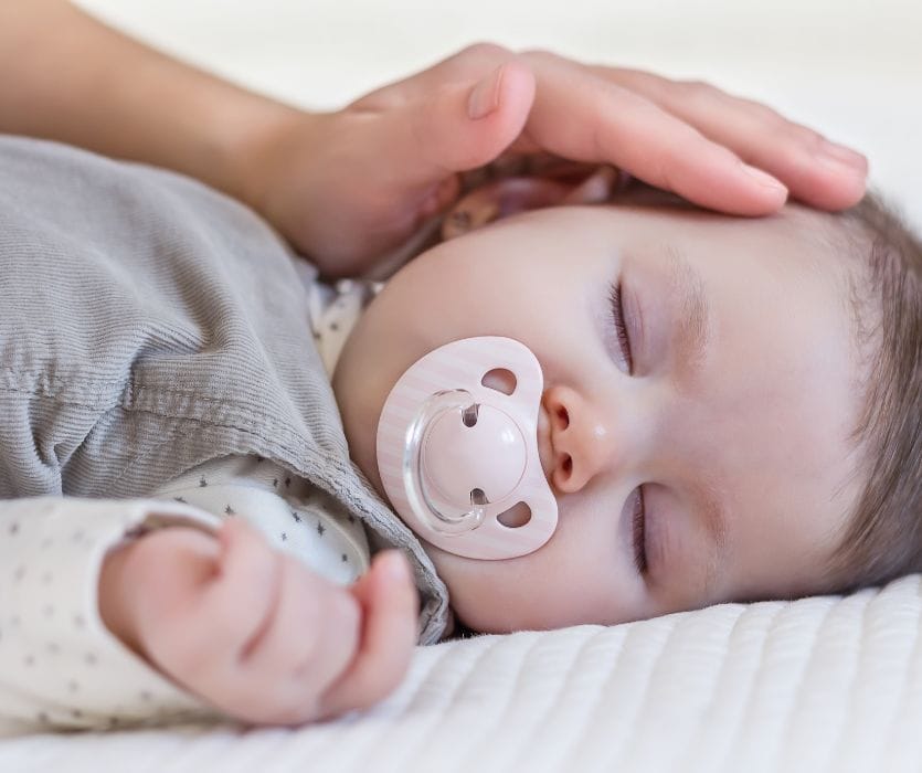 A sleeping infant with a pink pacifier rests on a white mattress while an adult hand gently strokes their head.