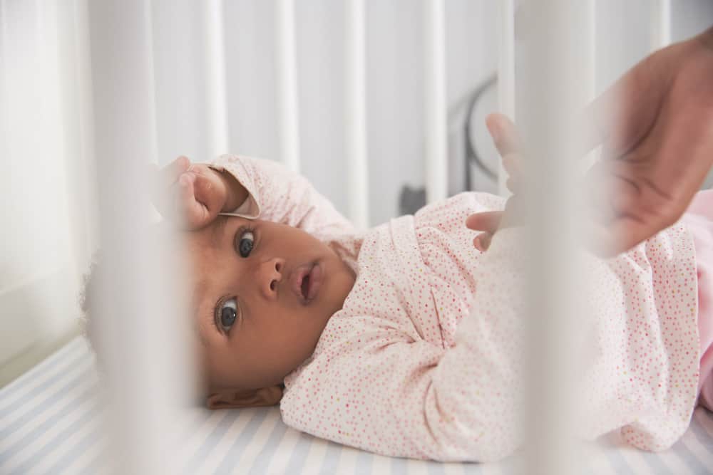 An infant lying in a crib looks up as an adult reaches a hand through the white slats toward them.
